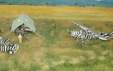 Miniatur-Flugzeug und Auto im Zebradesign in der Savanne. Ein Mann filmt, ein anderer steht neben einem Zelt. do 27.