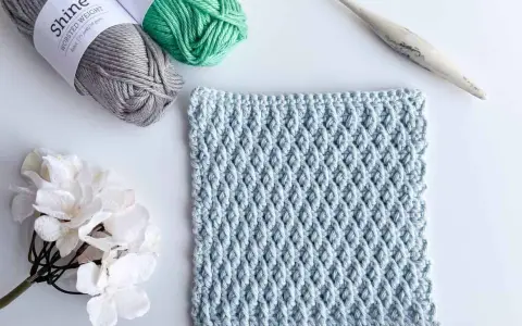 Muestra de tejido a crochet con el patrón nudo de salomon en color azul claro, junto a ovillos de lana gris y verde, una flor blanca y un ganchillo.