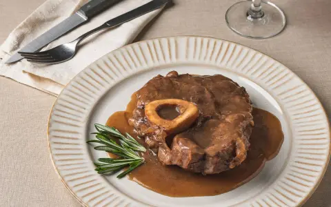 Un delicioso osso buco, tierno y jugoso, bañado en su salsa, servido en un plato con romero fresco.