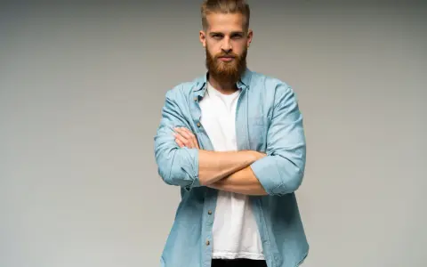 Uomo con barba e capelli biondi, camicia azzurra aperta su t-shirt bianca, braccia conserte. Look trasandato ma curato.
