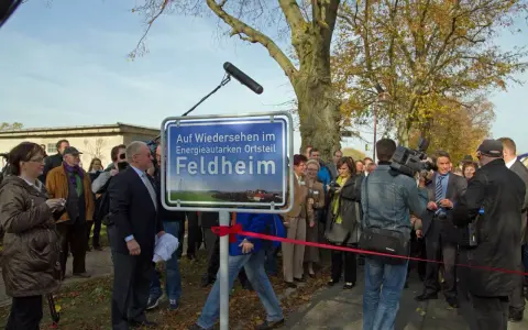 Feldheim, ein Vorreiter unter den energieautarken Gemeinden Deutschlands, wird hier mit einem Schild gefeiert. Eine Gruppe von Menschen, darunter ein Kameramann, versammelt sich.