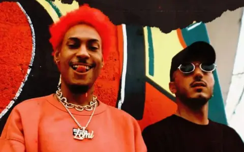 Dj Ballesteros y su colega posan frente a un mural vibrante. El primero, con cabello rojo y un collar dorado, sonríe con la lengua afuera.