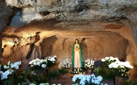 A statue of Mary stands in a grotto, surrounded by lush white flowers. The cave walls are illuminated, creating a serene atmosphere, reminiscent of the three fountains of grace.