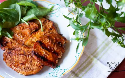 Grillowane plastry schabu w aromatycznej marynacie, podane z zielonymi liśćmi na talerzu.