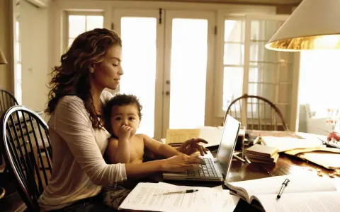 Una mamma lavoratrice con il suo bambino in grembo, mentre digita al computer tra libri e fogli.