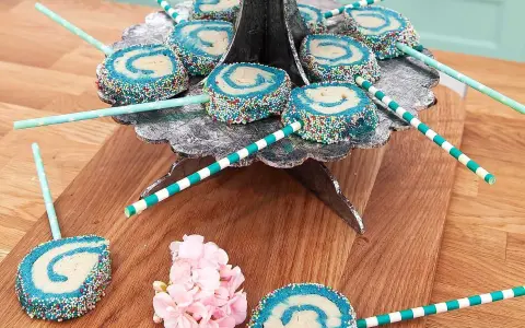 Blå och vita cake pops med strössel på ett silverfärgat fat. Vissa ligger på ett träunderlag med rosa blommor.
