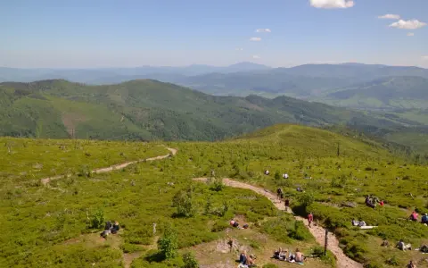 Malownicze widoki na Beskid Śląski, gdzie liczne istebne szlaki turystyczne prowadzą przez zielone wzgórza pełne jagód. Ludzie odpoczywają na trawie, ciesząc się słońcem.