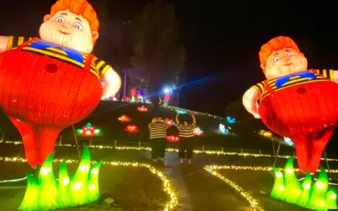 Gigantesche lanterne di bambini paffuti illuminano un sentiero a Wonderland Rimini, con due persone che salutano tra fiori luminosi.