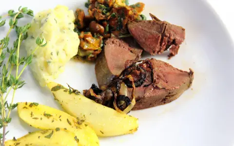 Rehleber zubereiten: zarte Scheiben Rehleber mit gebratenen Zwiebeln, Kartoffelpüree, gebratenen Kartoffelspalten und frischen Kräutern auf einem weißen Teller.