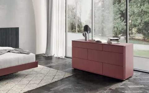 Cassettiera camera da letto moderna color bordeaux, con sei cassetti, accanto a un letto e una grande finestra.