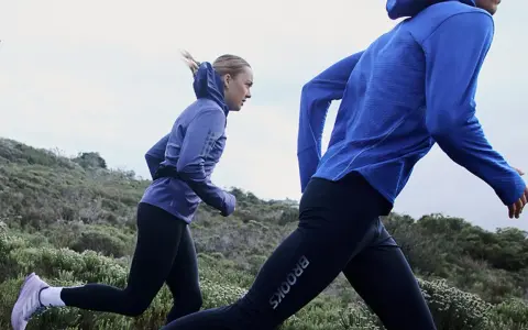 Två personer springer i en kuperad terräng. De är klädda i träningskläder och verkar inte bry sig om att det är lite grått ute, de njuter av att springa i regn.