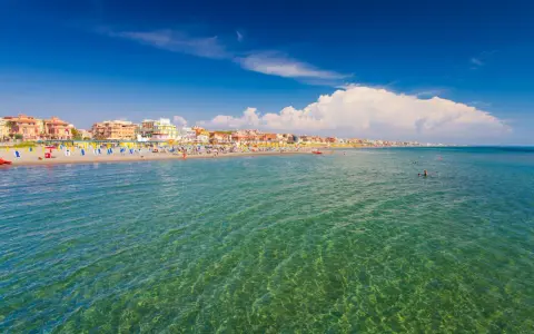 Słoneczna plaża w Rzymie, pełna kolorowych parasoli i ludzi kąpiących się w krystalicznie czystej wodzie.