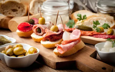 Pyszne tapas na drewnianej desce: pieczywo z wędliną, oliwkami, ogórkiem i suszonymi pomidorami.