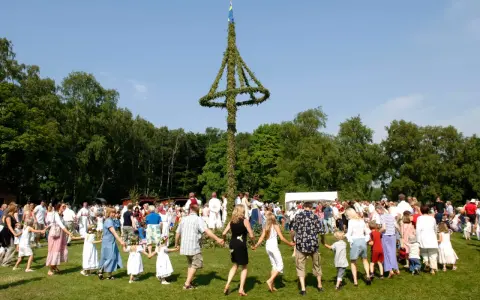 Människor dansar runt en midsommarstång i en grönskande park. Många barn med blomsterkransar i håret deltar i firandet.