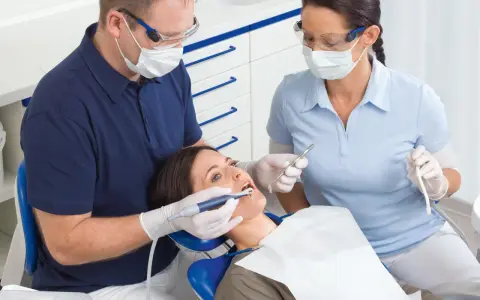 Dentysta i asystentka w okularach i maseczkach pracują przy pacjentce. Asystentka trzyma narzędzie, a dentysta używa **vector paro** do zabiegu.