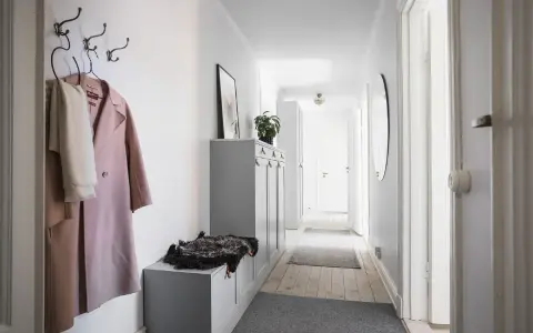 En smal hall med en rosa kappa och en vit halsduk på krokar. En grå förvaringsmöbel med en fårskinnspläd.