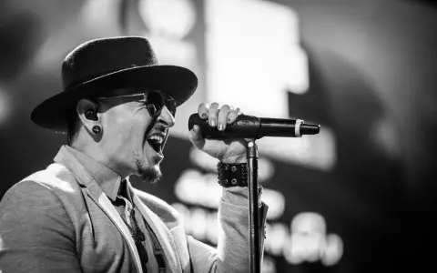 Wokalista Linkin Park, Chester Bennington, w kapeluszu i okularach przeciwsłonecznych, śpiewa do mikrofonu z pasją.