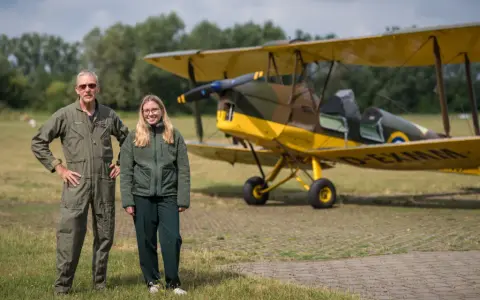 Mann in Fliegerkluft und junge Frau stehen neben gelbem Doppeldeckerflugzeug. Das Flugzeug erinnert an einen Tiger Moth.