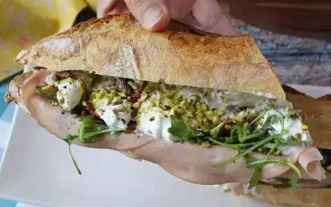 Bocadillo de pan rústico relleno de mortadela, burrata cremosa, rúcula, pistachos y tomates secos.