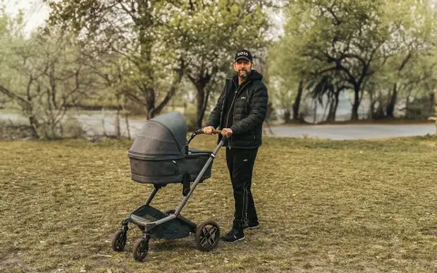 Mężczyzna w czapce z daszkiem i kurtce pcha nowoczesny wózek dla dziecka po trawie.