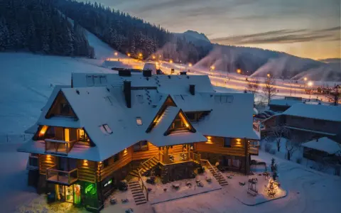 Drewniany pensjonat w śnieżnej scenerii, idealny na zakopane dla początkujących. Oświetlone stoki narciarskie w tle.