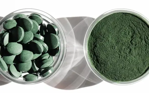 Dwie szklane miseczki: jedna z tabletkami chlorelli, druga z zielonym proszkiem. Dowiedz się, chlorella jak stosować, by czerpać z niej korzyści.