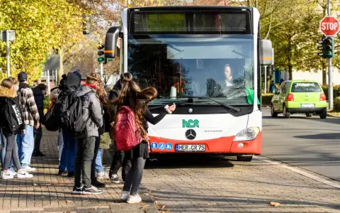 Ein Schulbus mit Warnblinker hält an einer Haltestelle. Schüler warten auf dem Gehweg, um einzusteigen.