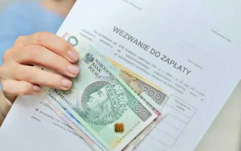 Dłoń trzyma wezwanie do zapłaty i polskie banknoty 100 i 200 zł. Dokument zawiera informację o zawiadomieniu o uchyleniu zajęcia.