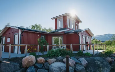 Rött hus med vita knutar och en solig veranda. Abisko Turiststation välkomnar besökare till fjällen.