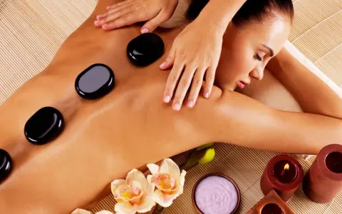 A woman enjoys the benefits of hot stone massage, with smooth black stones placed on her back. Orchids and candles add to the serene spa atmosphere.