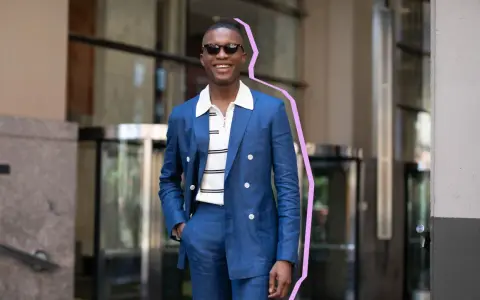 Hombre sonriente con traje azul y camiseta blanca a rayas, gafas de sol, posando con la mano en el bolsillo.