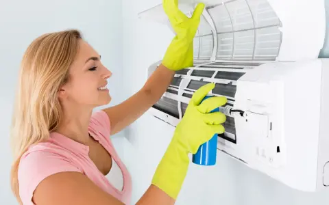 Mujer con guantes verdes limpia el aire acondicionado que huele a humedad, rociando un producto.