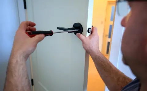 Un homme utilise un tournevis pour fixer une poignée de porte noire. Il est sur le point de lever la poignée pour fermer la porte.