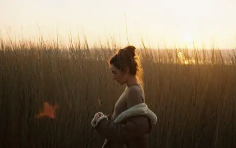Une jeune femme, le regard baissé, se tient dans les hautes herbes au crépuscule. La lumière dorée du soleil couchant illumine ses cheveux et le paysage.