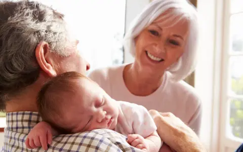 Los abuelos paternos sonríen mientras el abuelo sostiene al bebé dormido en su hombro.