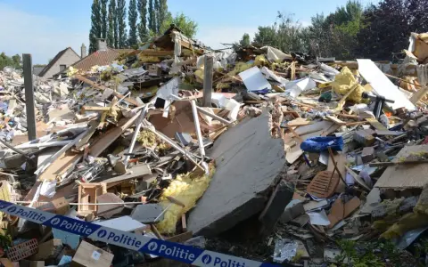 Ogromna kupa gruzów po zawaleniu się budynku. Widać izolację, drewno, beton i niebieską torbę. Wstęga policyjna zabezpiecza teren.