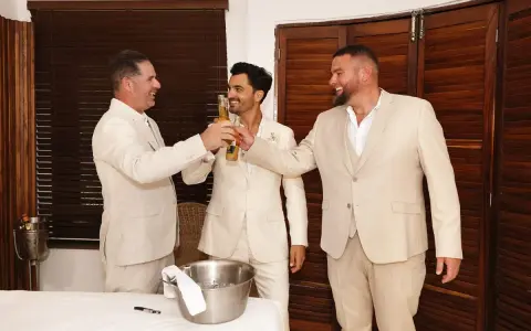 Tres hombres en trajes claros brindan con cerveza. El novio, en el centro, sonríe. Un momento de alegría antes del discurso de boda hermano.
