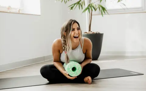 Lachende Frau mit Faszienrolle auf Yogamatte. Sie strahlt Lebensfreude aus und zeigt, wie man das positive sehen kann, auch nach dem Training.