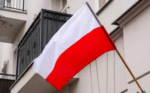 Flaga Polski w pionie, biało-czerwona, powiewa na tle budynku z balkonem.