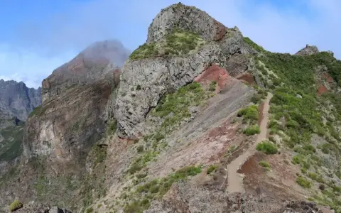 Steiler Bergpfad auf Madeira, ideal für die Packliste. Felsige Gipfel, grüne Vegetation und ein Weg, der sich durch die zerklüftete Landschaft schlängelt.