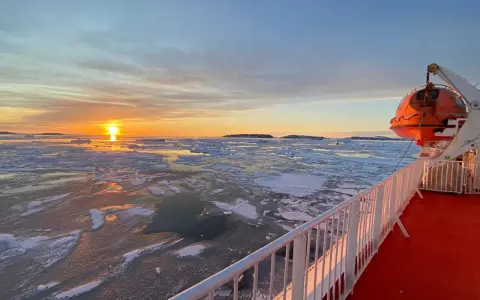A **Greenland ferry** navigates icy waters at sunset. An orange lifeboat hangs ready on the ship's deck.