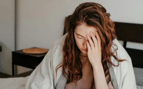 Kvinna med rött hår i sängen, täckt av filt, håller handen för ansiktet. Hon ser ut att ha feber, ett kolera symptom.
