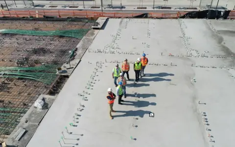 Grupa budowlańców na placu budowy, gdzie trwa wylewanie betonu. Zastanawiają się, ile kosztuje gruszka betonu, analizując postęp prac.