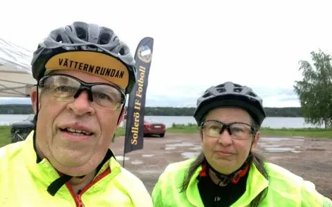 Två cyklister redo för Vätternrundan. Mannen har en keps med texten "Vätternrundan". Bakom dem syns en sjö och en flagga för Sollers IF Fotboll.