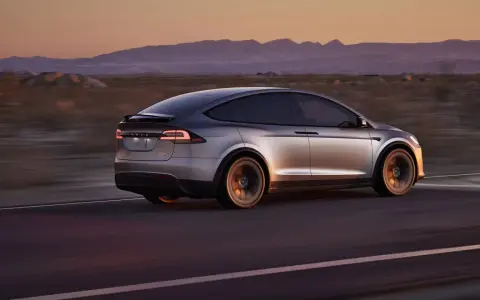 Srebrny Tesla X pędzi przez pustynny krajobraz o zachodzie słońca. Zastanawiasz się nad jego ceną?
