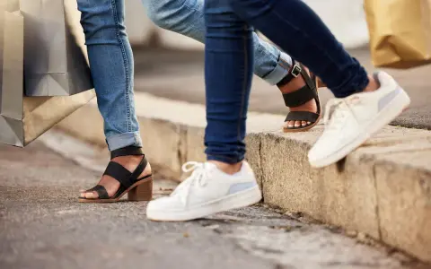 Dwie osoby w jeansach i modnych butach, z torbami na zakupy w rękach, idą chodnikiem. Jedna ma czarne sandały na obcasie, druga białe sneakersy.