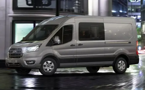 Camión N1 gris Ford Transit circulando por una calle urbana mojada de noche, con luces de edificios y tráfico difuminadas al fondo.