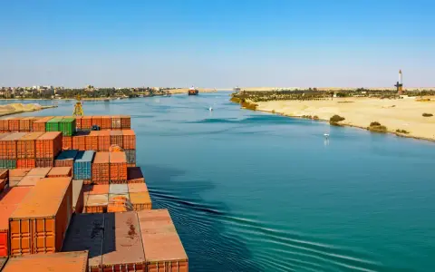 Containerrieschiff fährt durch den Suezkanal, ein Meisterwerk des Suezkanal Baus, vorbei an Wüstenlandschaft und Stadt.