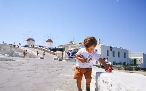Un bambino gioca con una macchinina rossa a Mykonos, con i mulini a vento sullo sfondo. Vacanze a Mykonos con bambini.