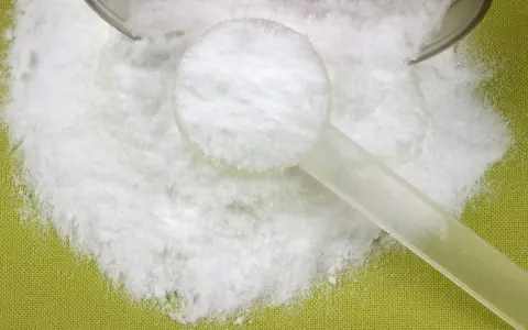 Biały proszek, prawdopodobnie l-glutamina, rozsypany na zielonym tle, z plastikową miarką.